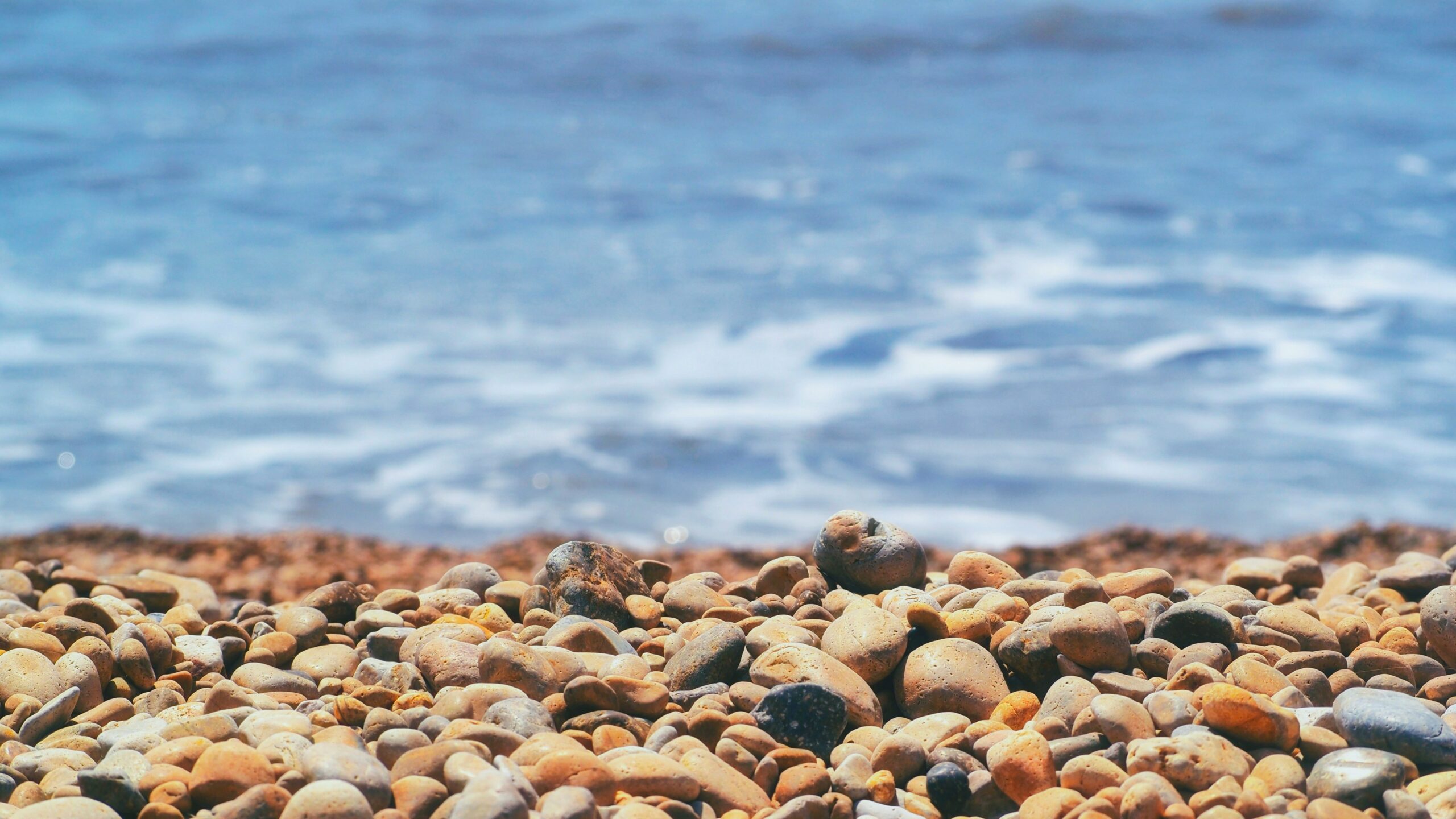 Premier plan lumineux sur une plage de galets lisses et dorés face à une mer bleue apaisante, symbolisant l'ancrage, la sérénité et le chemin vers le bien-être chez Un Souffle d’Art-Mot-Ni.